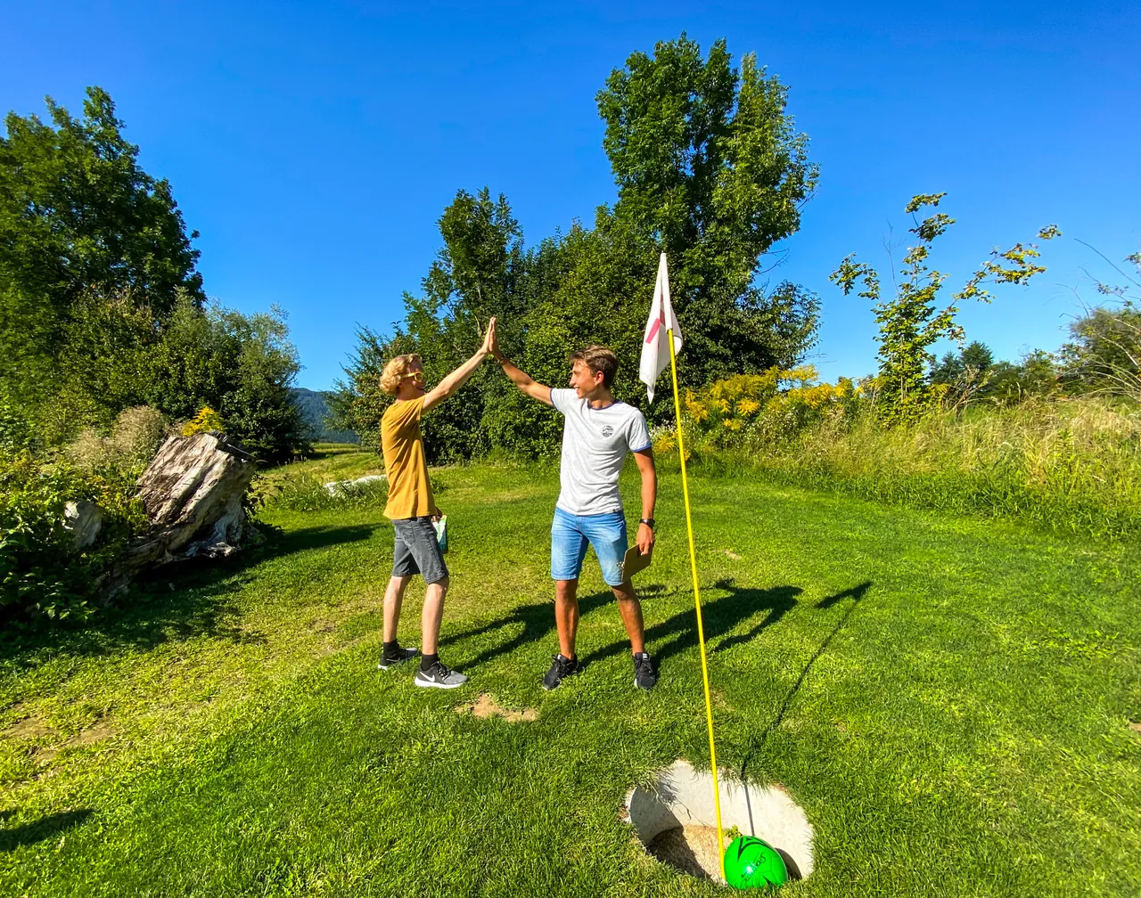 Zwei Spieler klatschen sich am Fußballgolf-Loch ab; der grüne Ball liegt im Ziel